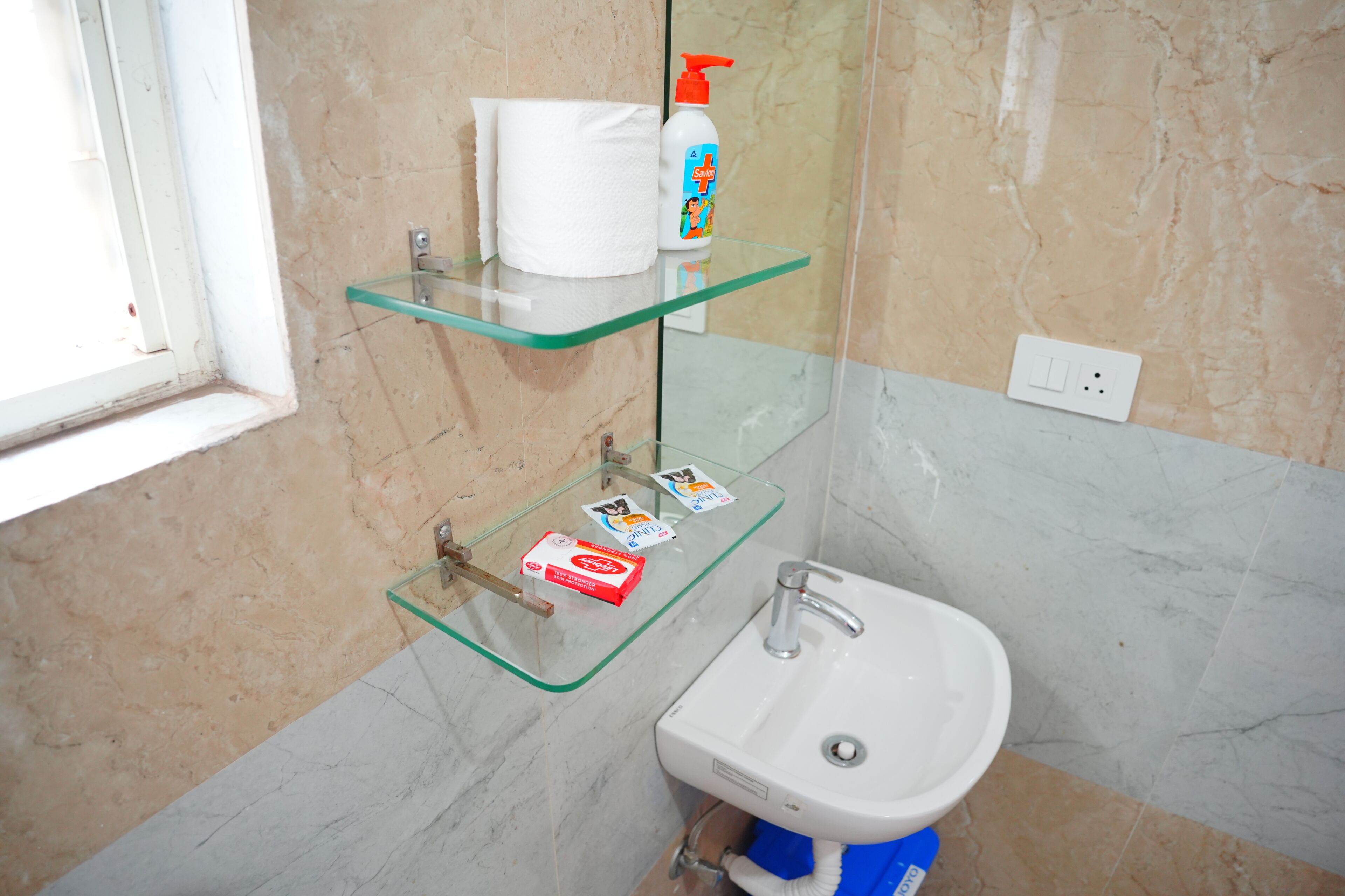 Baño con repisa de vidrio que sostiene un rollo de pañuelos, una botella de jabón Savlon y una caja de jabón Dove.