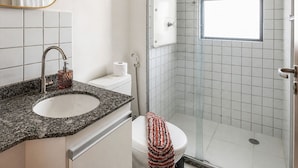 A bathroom with a granite countertop sink, a toilet, a shower with a glass door, and a window providing natural light. - CJC Short Stay Ecogarden (Natal)