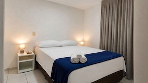 A hotel room with a bed, two bedside lamps, a nightstand, a rolled towel on the bed, and a curtain. - CJC Short Stay Ecogarden (Natal)