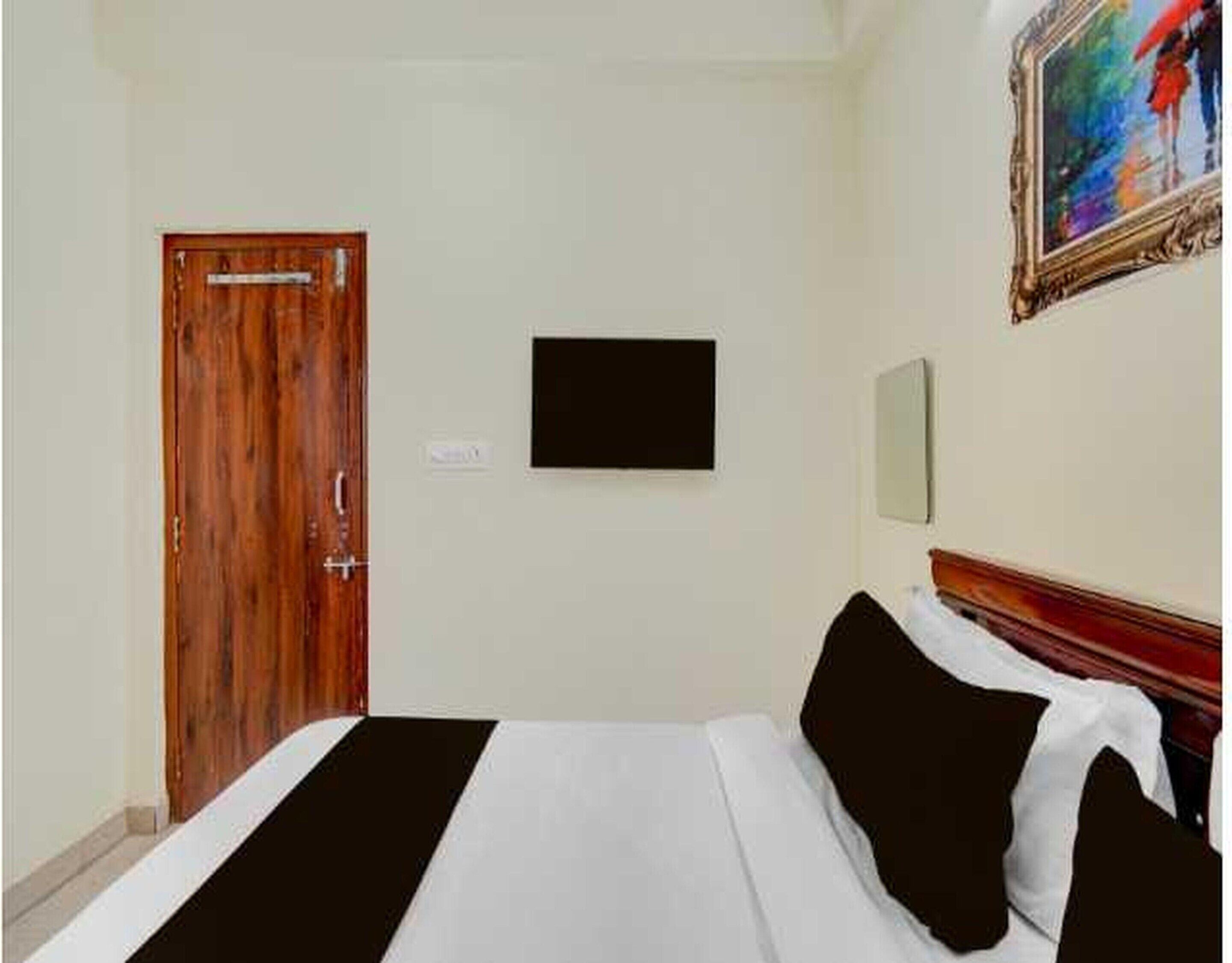 A hotel room with a wooden door, a bed with black and white bedding, a flat-screen TV mounted on the wall, and a painting above the bed.