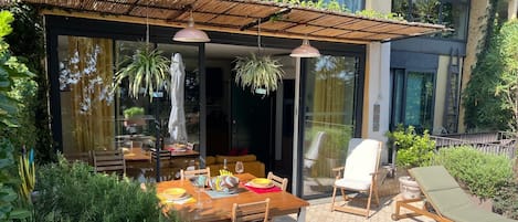Un espace de restauration en plein air avec une table et des chaises en bois, un fauteuil confortable et une pergola ornée de plantes suspendues.