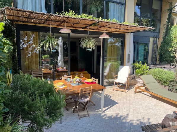 An outdoor dining area with a wooden table and chairs, a cushioned lounge chair, and a pergola with hanging plants.