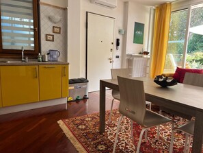 A compact kitchen with yellow cabinets, a dining table with chairs, a patterned rug, and a window with yellow curtains. - Lake of Varese, "Residenza Mirabella" (Gavirate)