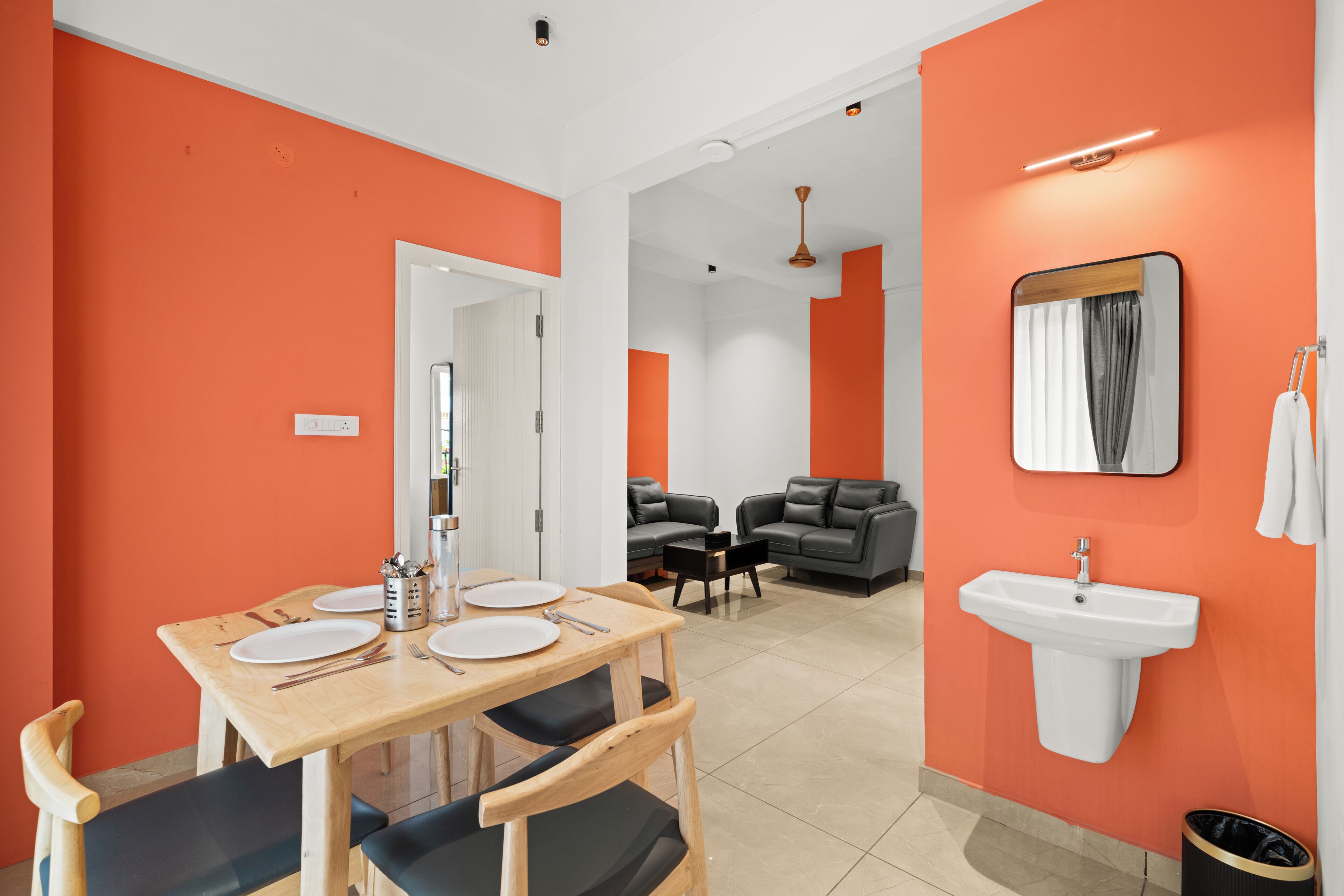 A modern hotel room with orange walls, a dining area with a wooden table and chairs, a sofa, a small black sofa, a white sink, and a mirror.