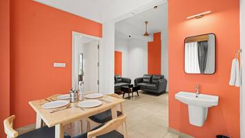A modern hotel room with orange walls, a dining area with a wooden table and chairs, a sofa, a small black sofa, a white sink, and a mirror.
