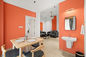 A modern hotel room with orange walls, a dining area with a wooden table and chairs, a sofa, a small black sofa, a white sink, and a mirror.