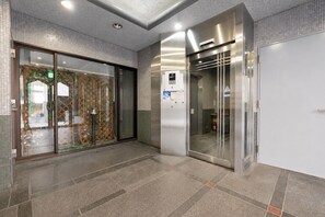 A modern elevator with a control panel and emergency button, a glass door with a patterned design, and a tiled floor. - b&Dotonbori 1 (Chuo Ward)