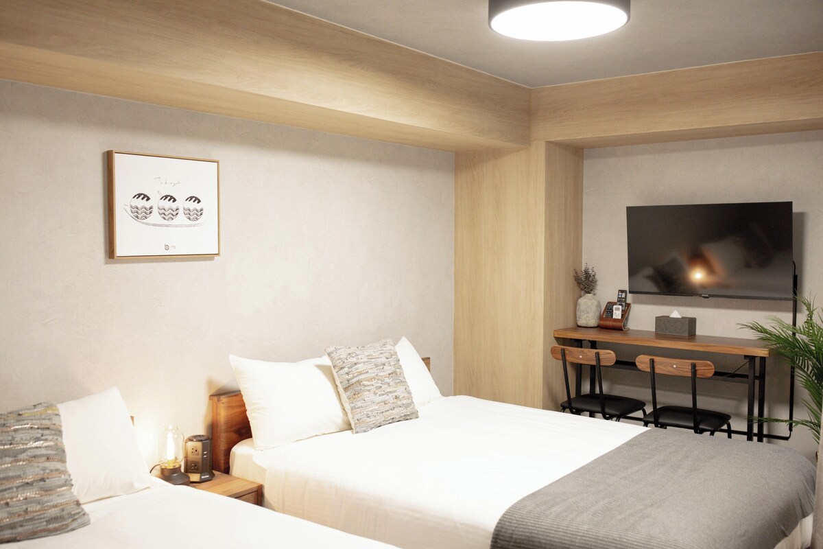 A hotel room with a bed, bedside tables, a desk with chairs, a television, and a framed picture on the wall.