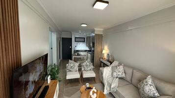 Uma sala de estar moderna com televisão de tela plana, um sofá, duas poltronas, uma mesa de centro e uma área de cozinha ao fundo.