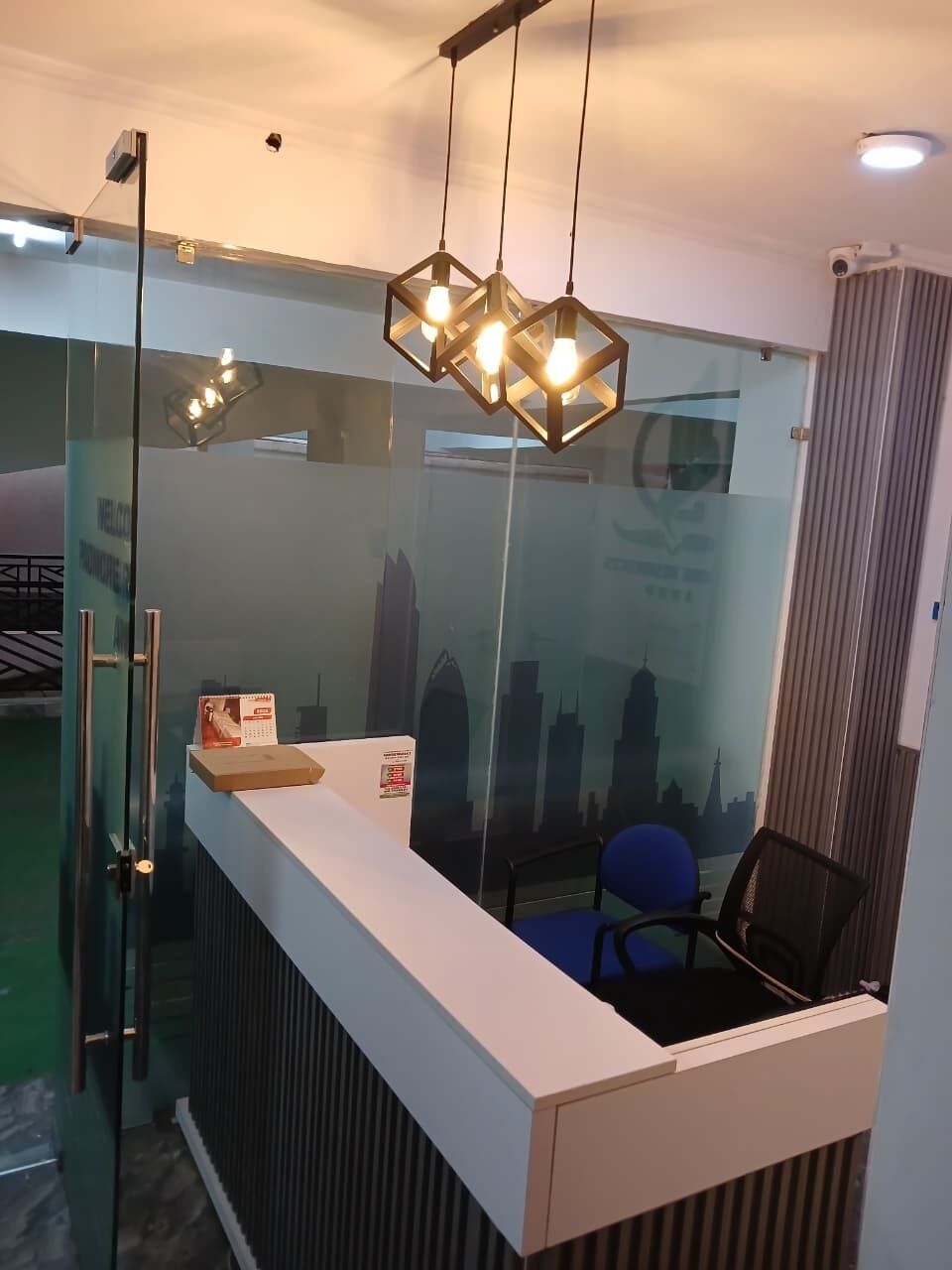 Un mostrador de recepción con una partición de vidrio, una silla azul y un cartel con un paisaje urbano.