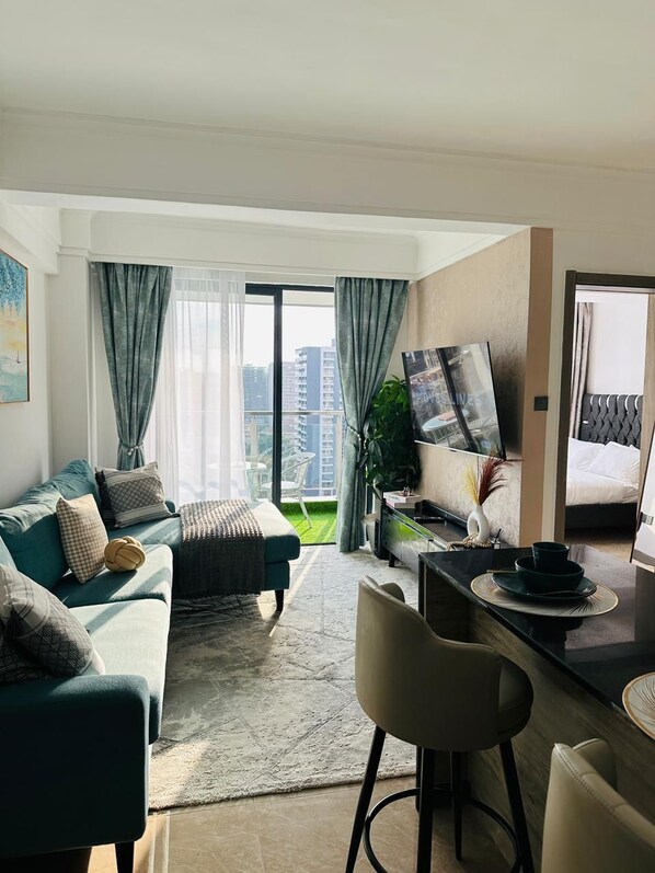 A modern living room with a sofa, a dining area with a table and chairs, a TV mounted on the wall, and a balcony with a view of the city. - Padmore Residences Kilimani (Nairobi)