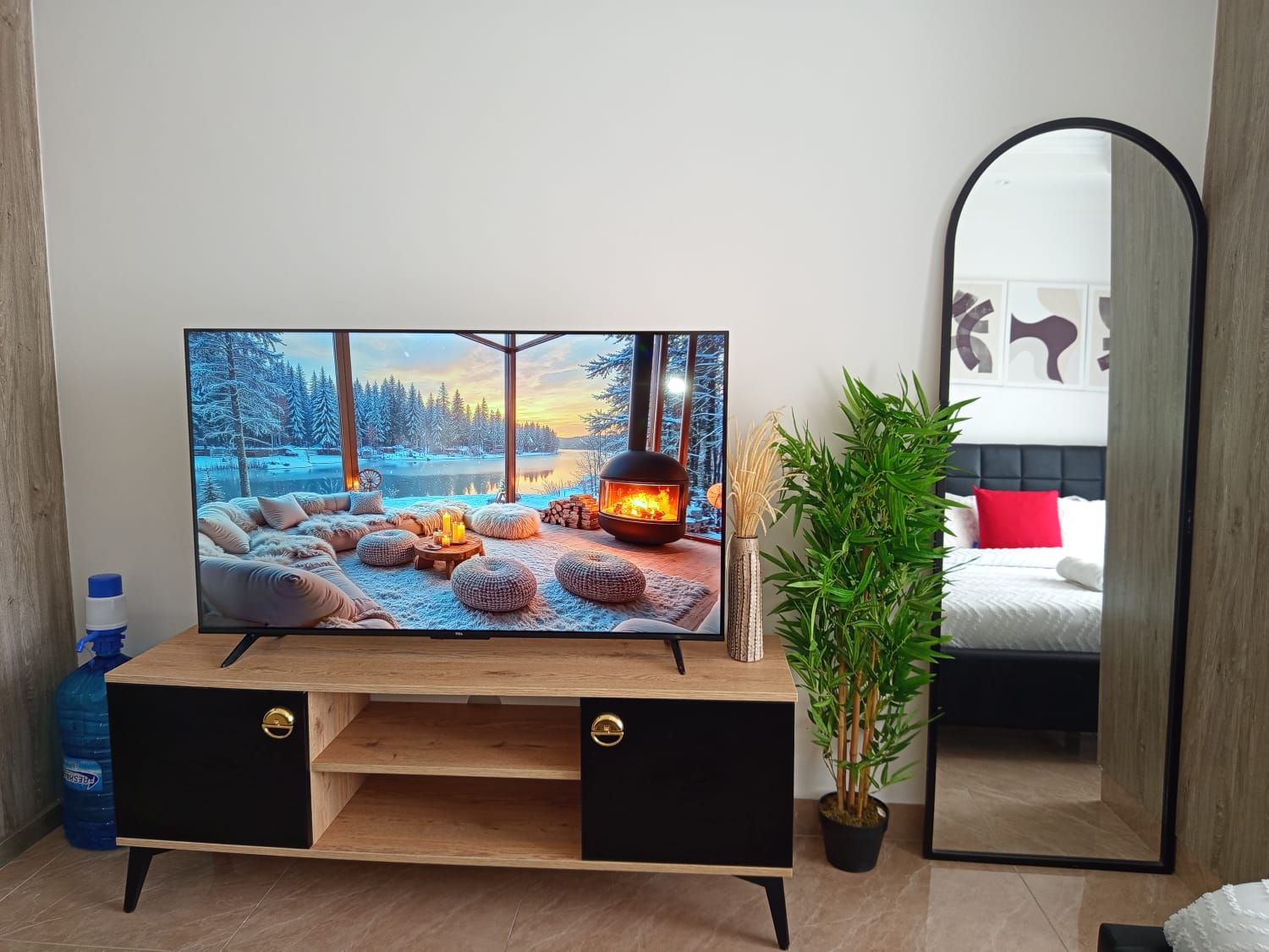 A modern living room with a large flat-screen TV, a wooden entertainment unit, a potted plant, and a bed with a red pillow.
