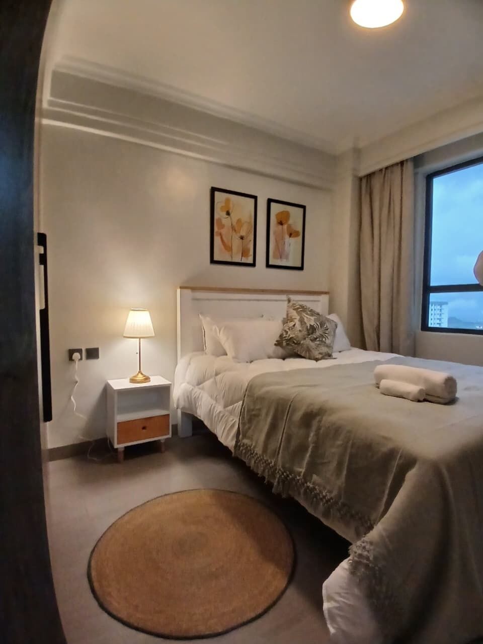 A hotel room with a bed, bedside table, lamp, artwork on the wall, and a view of the outside.