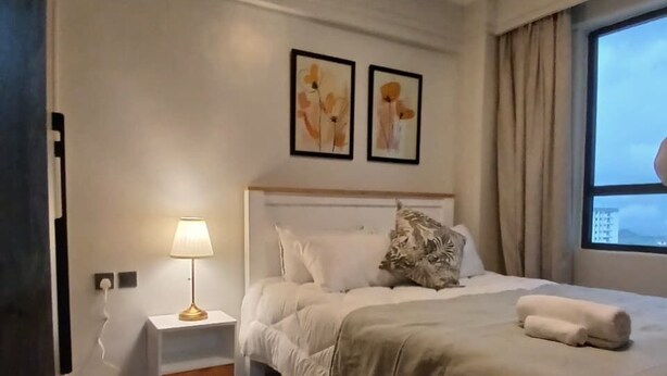 A hotel room with a bed, bedside table, lamp, artwork on the wall, and a view of the outside. - Padmore Residences Kilimani (Nairobi)