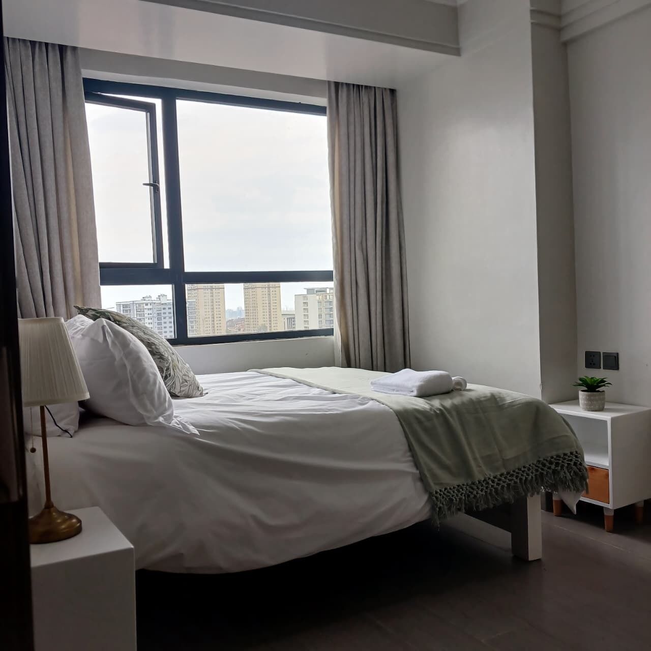 A modern hotel room with a large window, a bed with white linens, a bedside table with a lamp, and a small plant on a nightstand.