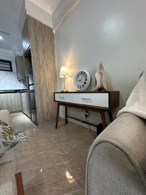 Una sala de estar con sofá, una mesita de noche con lámpara, un reloj y un estante en la pared.