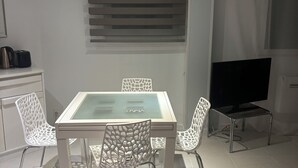 A modern dining area with a glass-top table, white chairs with intricate cut-out patterns, a flat-screen TV, and a kitchen area with a kettle and toaster. - Vacation Apartment (La Croix-Valmer)