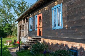 Ein Holzhaus mit roter Tür und blauen Fensterrahmen, eine Treppe führt zum Eingang, und ein Fundament aus Ziegelsteinen.