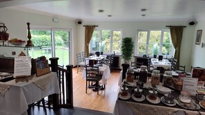 A breakfast area with tables set for dining, a sign detailing the menu, and a view of the outdoors through large windows. - Dolbrodmaeth Riverside  (Machynlleth)