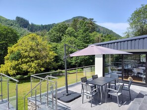 Una terraza con mobiliario exterior, barandilla de cristal y vista a una ladera boscosa.