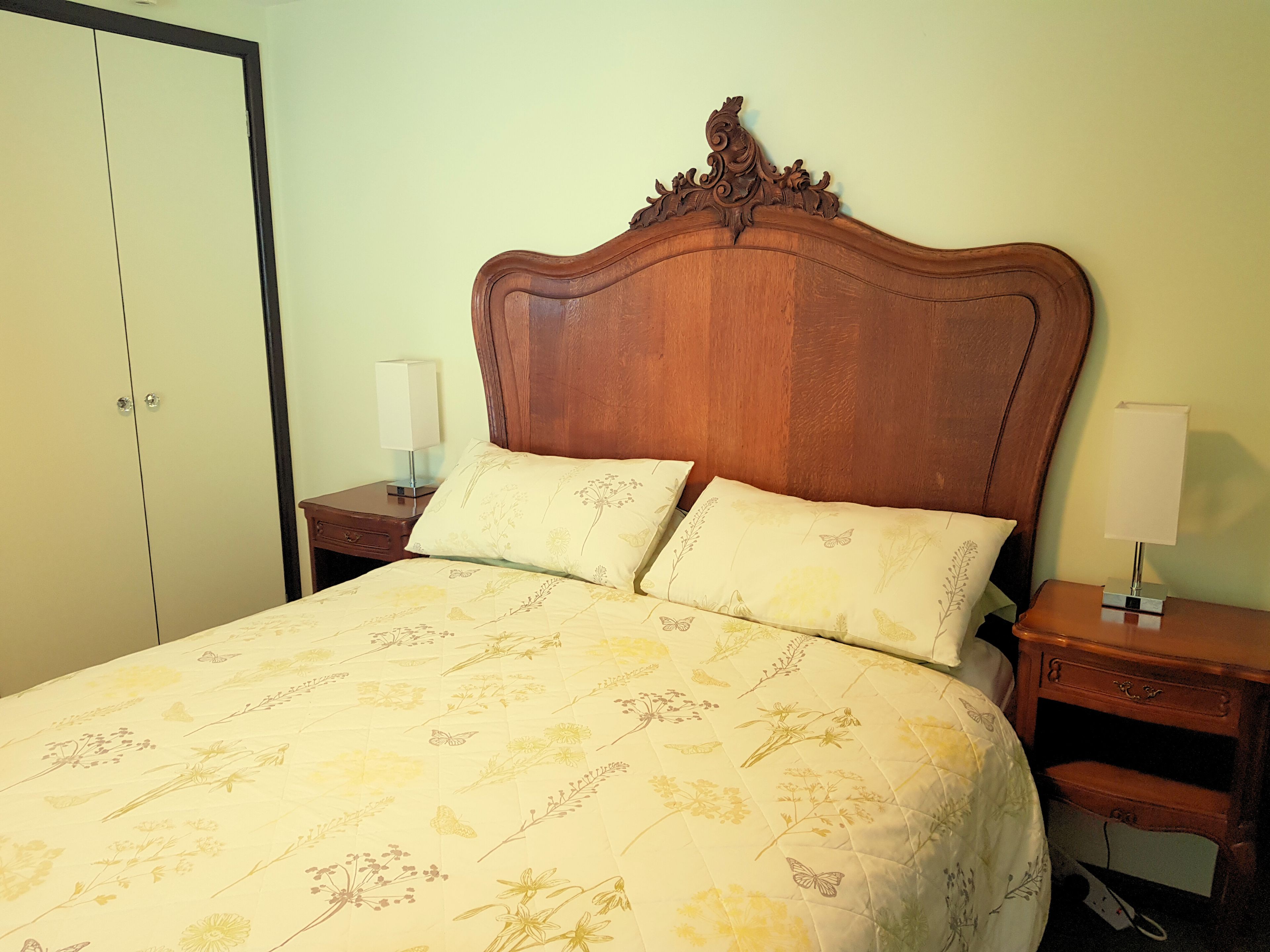 Una cama con una colcha de estampado floral, un cabecero de madera con detalles tallados, dos mesitas de noche con lámparas y un armario con estructura negra.