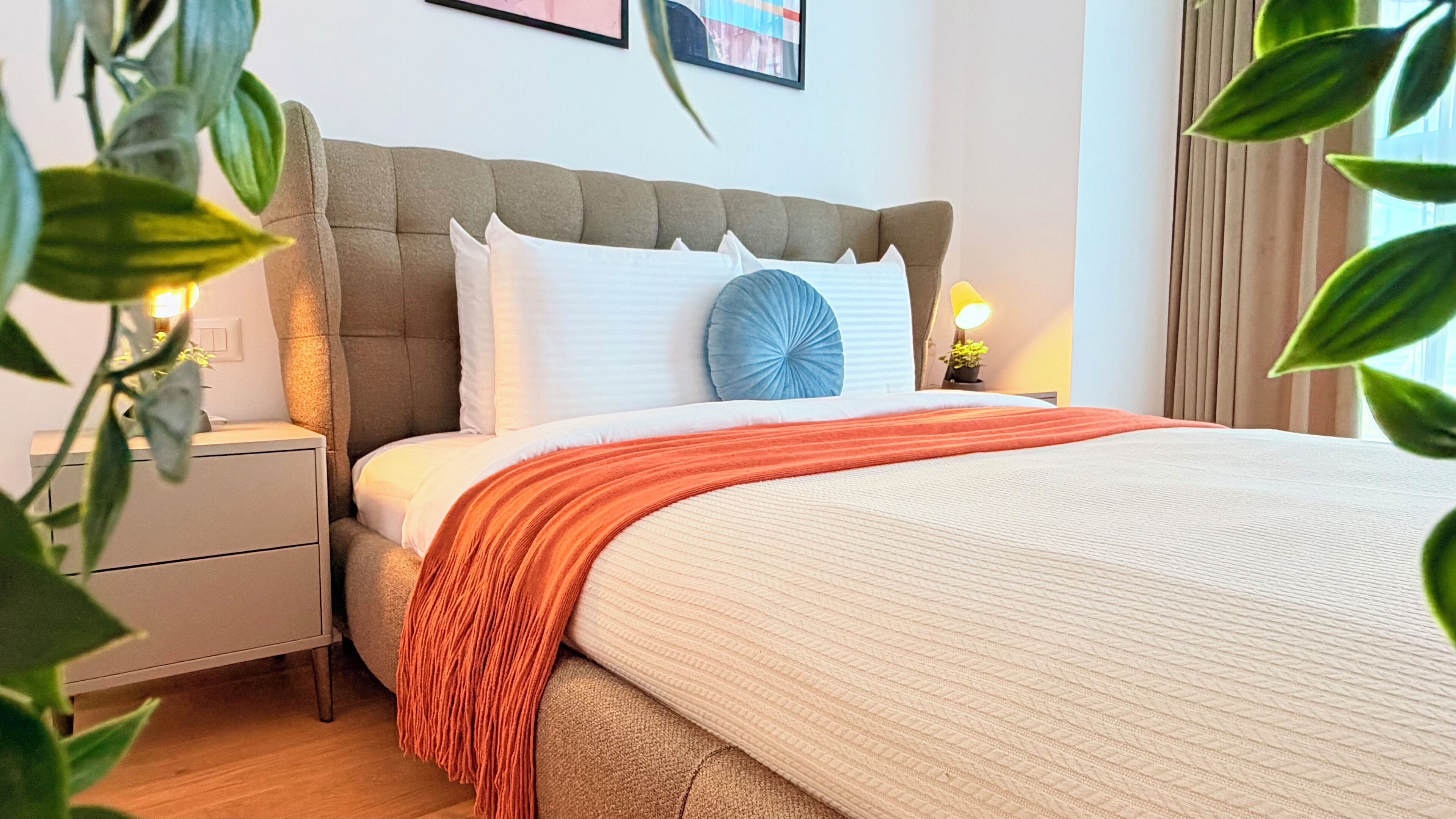 Uma cama bem arrumada com um cobertor branco e laranja, uma cabeceira estofada cinza e uma mesa de cabeceira com abajur.