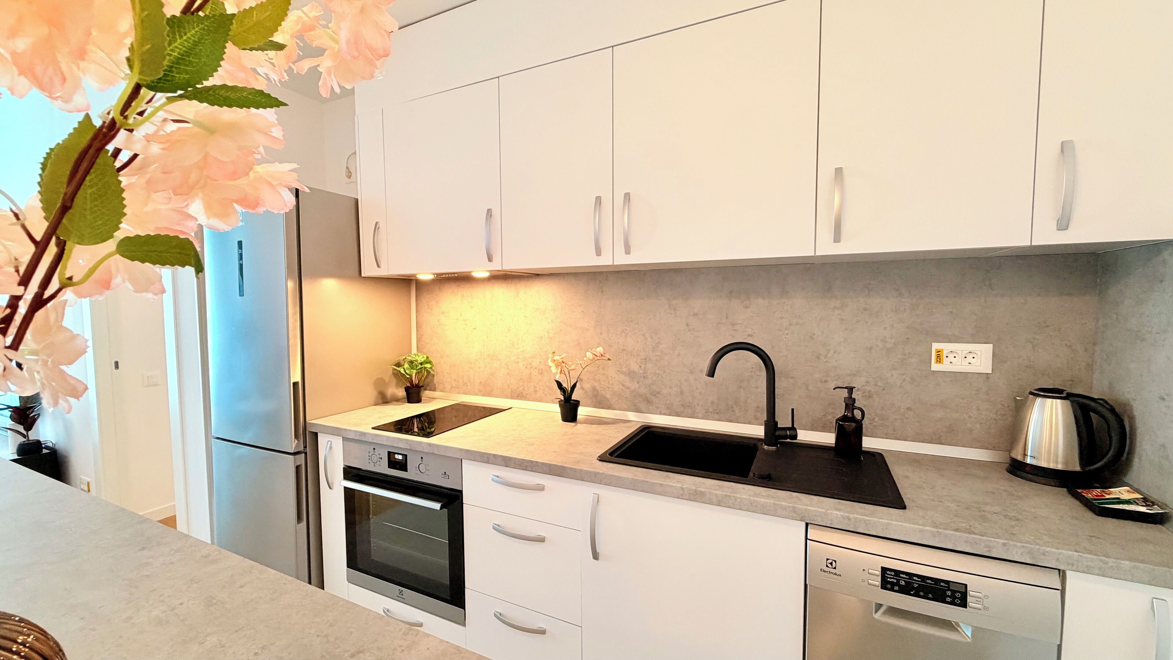 Una cocina moderna con armarios blancos, un refrigerador de acero inoxidable, un horno, un lavavajillas, un lavamanos con grifo negro y una tetera sobre la encimera.