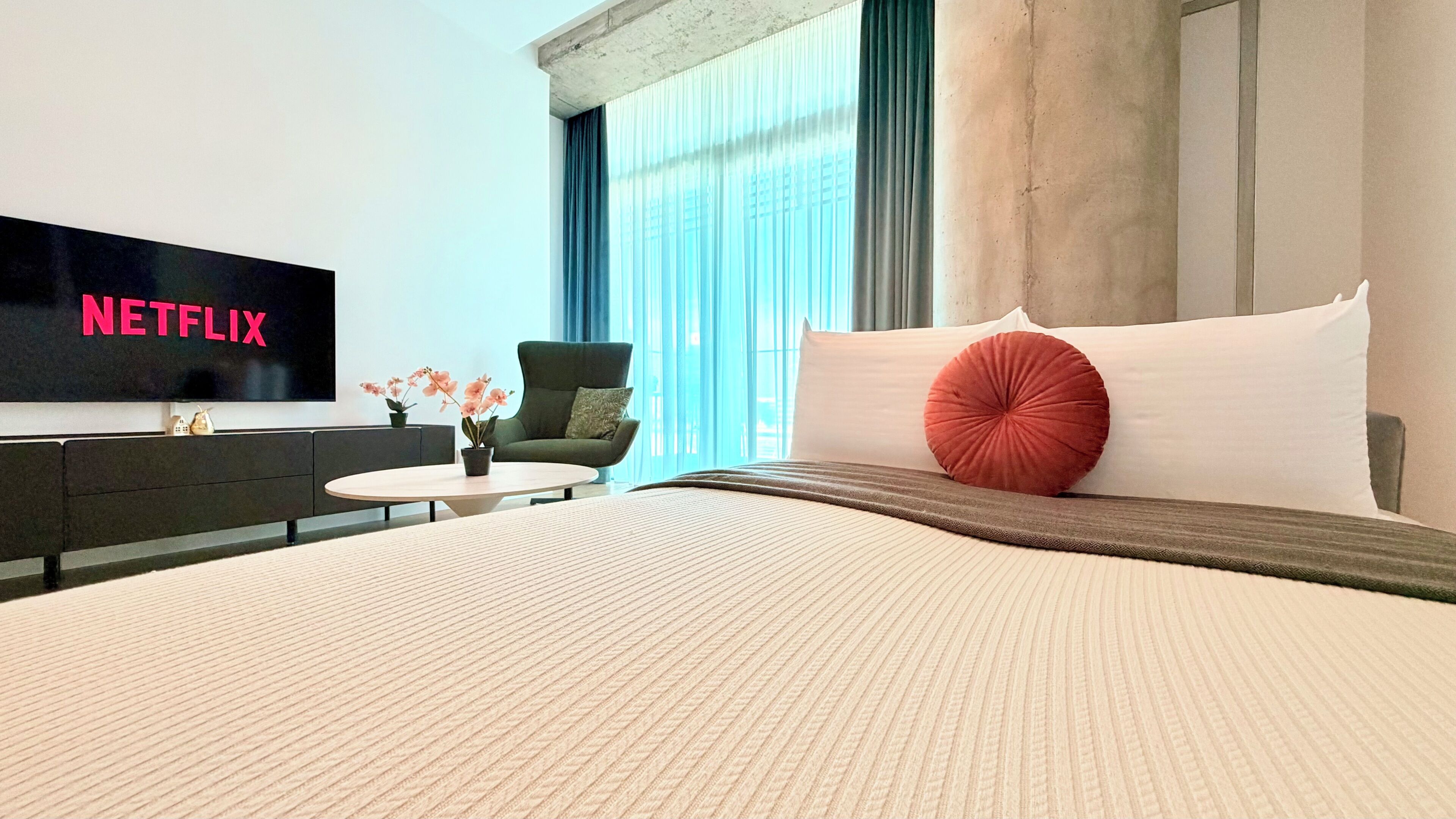 Habitación de hotel con una cama grande, un televisor de pantalla plana mostrando 'NETFLIX', una mesita redonda con un florero y un sofá.