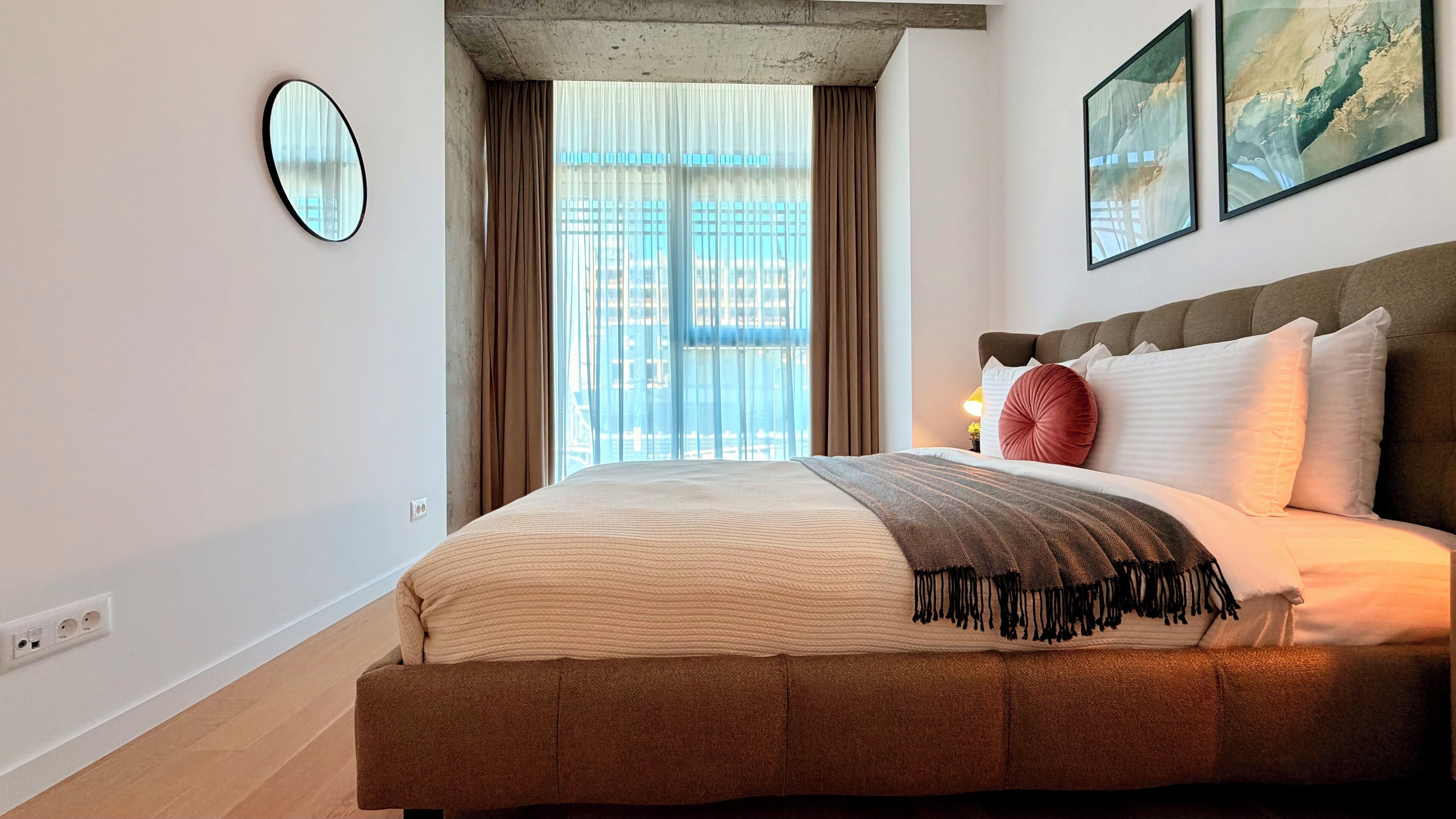 Ein Schlafzimmer mit einem großen Bett, einem runden Spiegel, einem Fenster mit Vorhängen und zwei gerahmten Bildern an der Wand.
