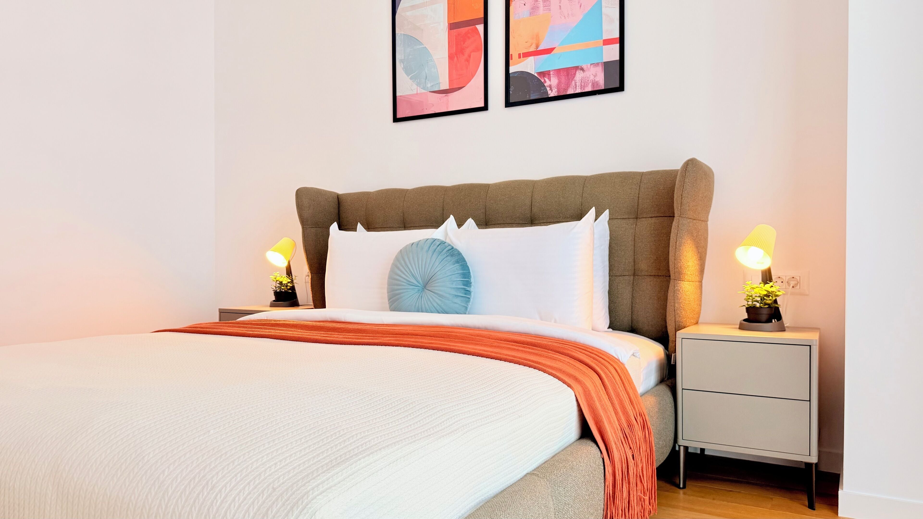 Uma cama bem arrumada com edredom branco e manta laranja, com duas mesas de cabeceira com luminárias e plantas em vasos.