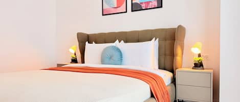 A neatly made bed with a white comforter and orange throw blanket, flanked by two bedside tables with lamps and potted plants.