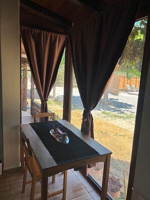 A room with a table and chairs, a window with curtains, and a view of an outdoor area with a fence and chairs. - Papyrise Nest (Heraklion)