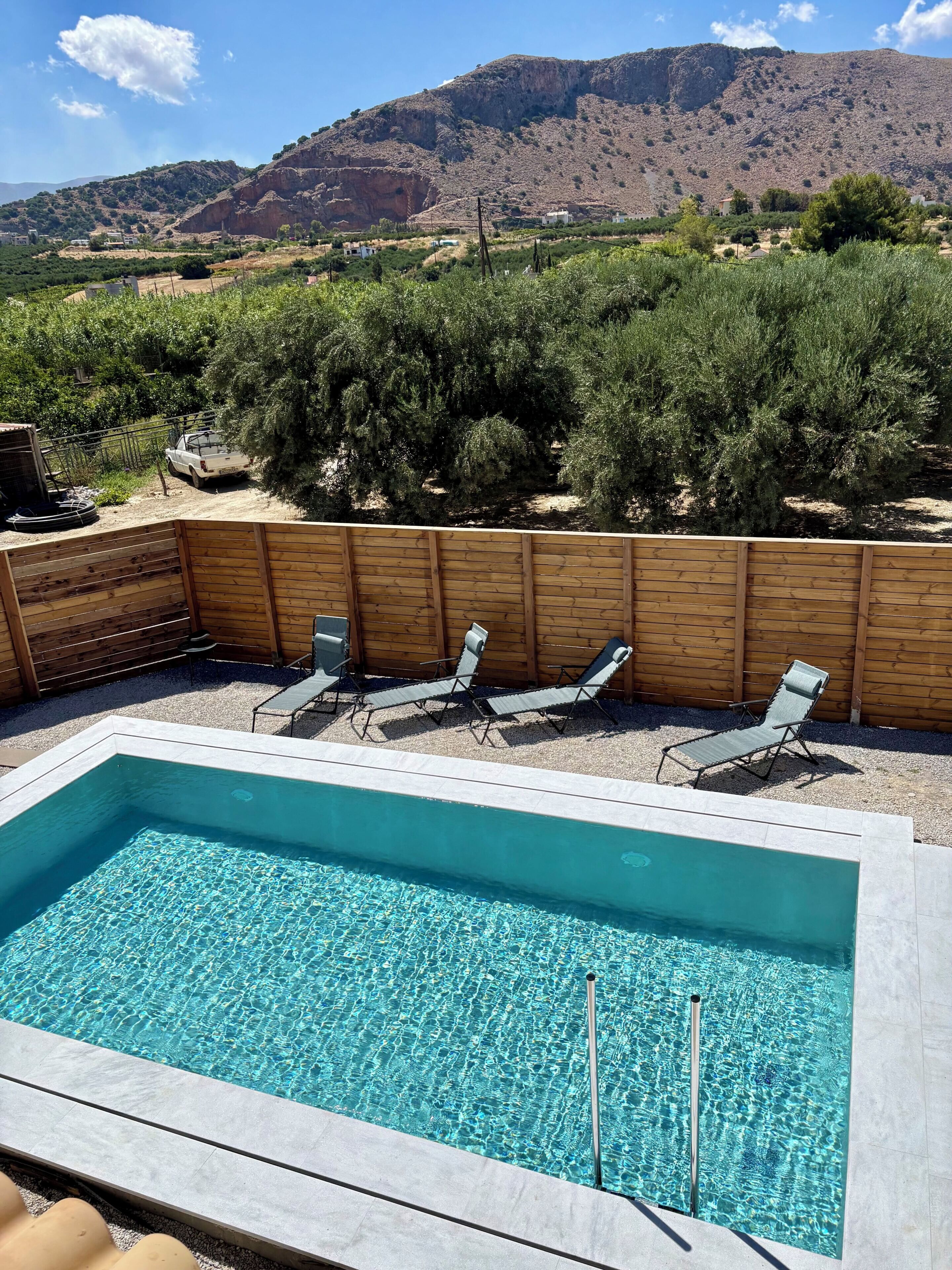 Una piscina rectangular con agua cristalina, rodeada por una valla de madera, y tres sillas de descanso.