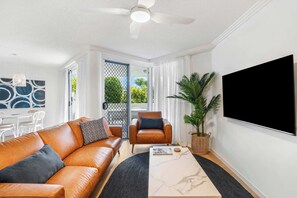 A modern living room with a leather sofa, a coffee table, a TV, and a large window with curtains. - Ocean Vista On Alex Unit 4 (Alexandra Headland)