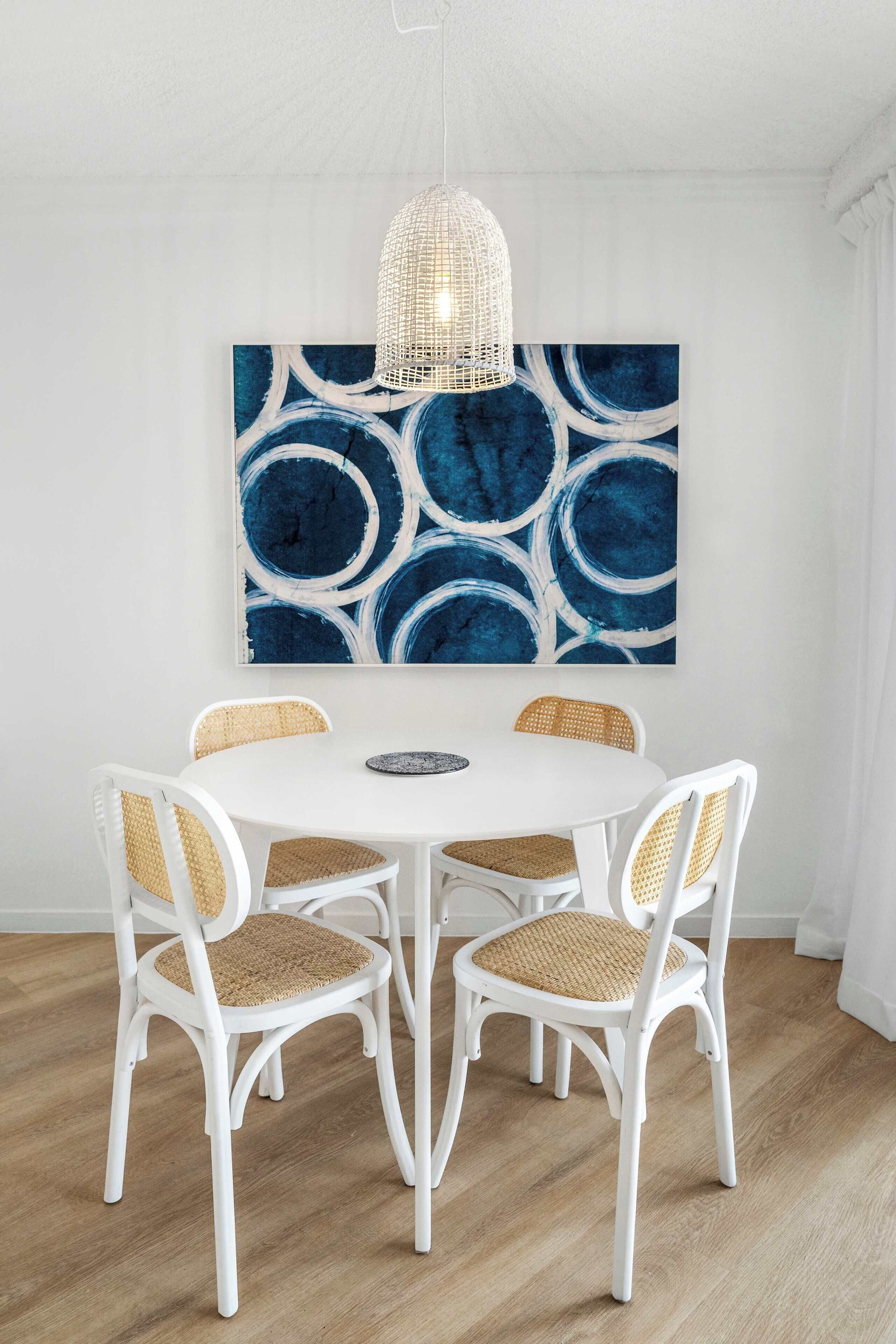 A dining area with a white round table surrounded by four wicker chairs, a woven pendant light, and a large abstract blue painting on the wall.