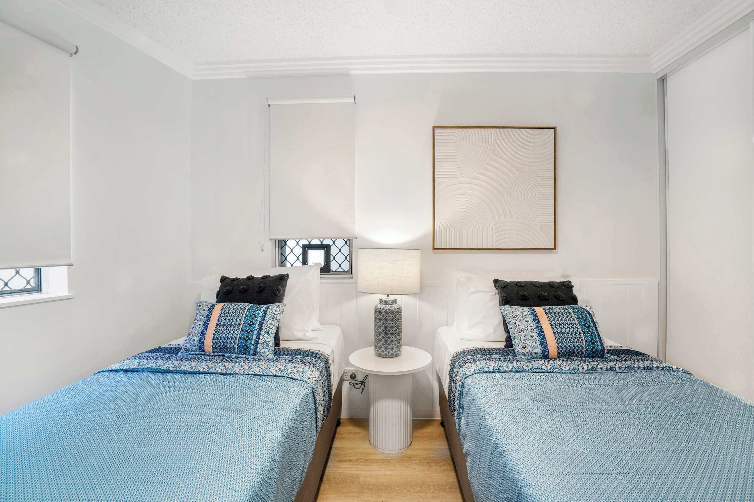 A hotel room with two beds, a small table, a lamp, and a framed picture on the wall.