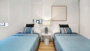 Hotellihuone, jossa on kaksi sänkyä, pieni pöytä, lamppu ja seinällä oleva kehystetty kuva.