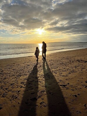 A beach scene with footprints leading towards the water, a person and a child walking, and the sun setting in the background.