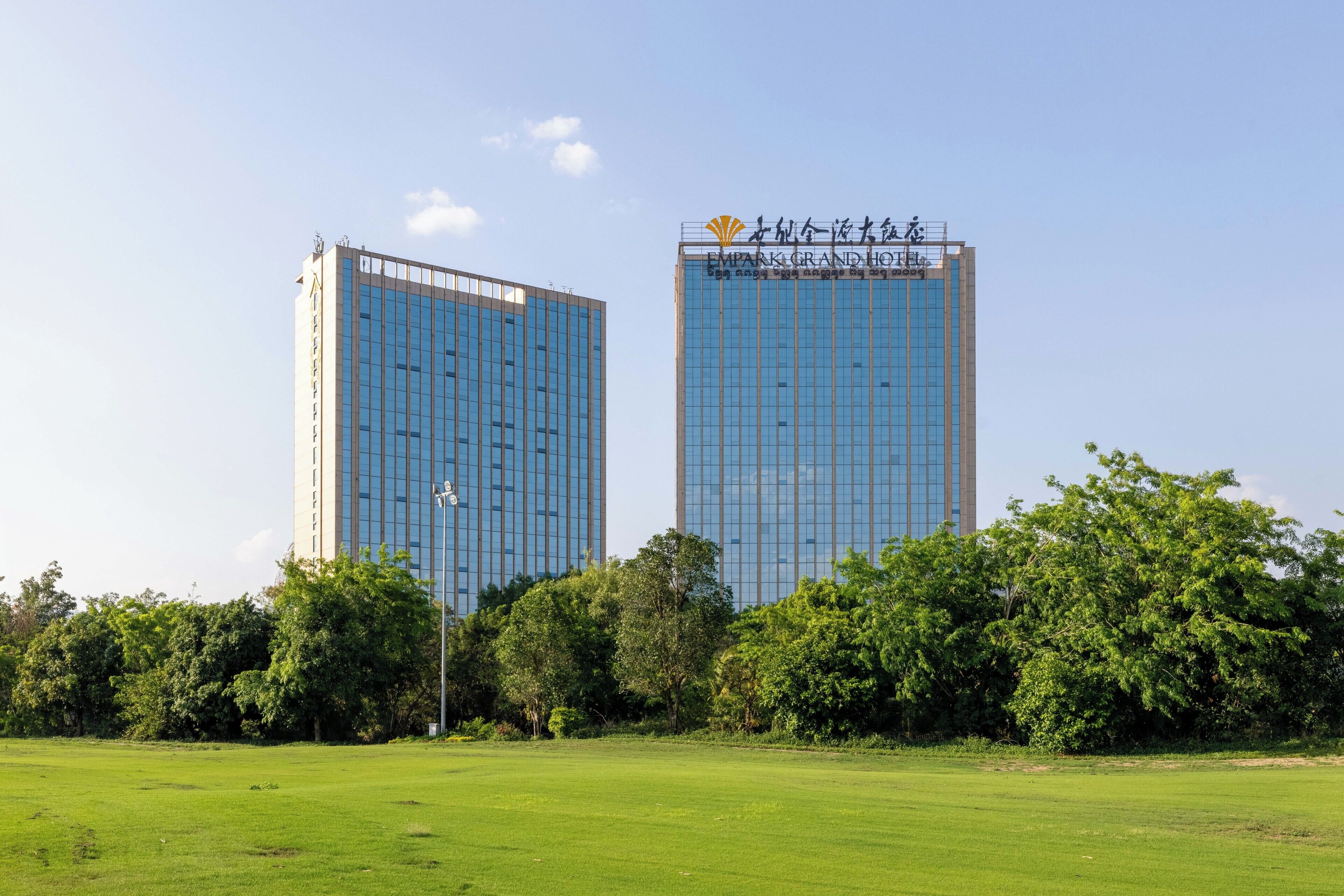 Two high-rise buildings with reflective glass facades, surrounded by a grassy field and dense trees.