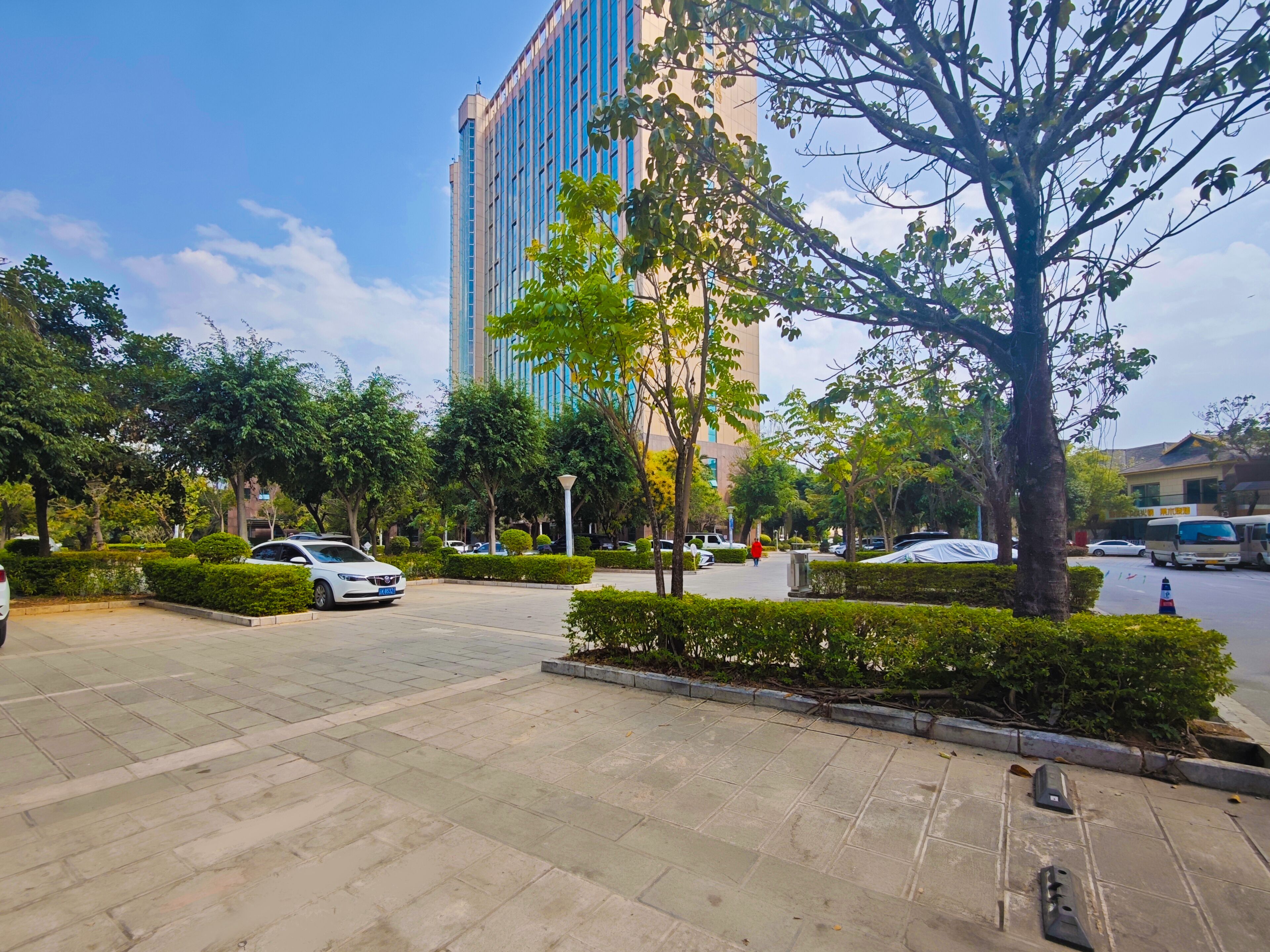 A paved outdoor area with a car, trees, and a tall building in the background.