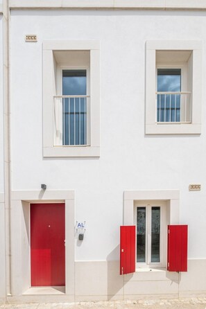 Un bâtiment avec une porte rouge, deux fenêtres à cadres blancs et une petite fenêtre avec des volets rouges.