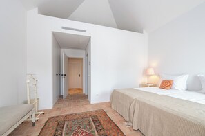 Une chambre à coucher comprenant un lit, une table de chevet, une lampe, une commode, un porte-vêtements suspendu, un tapis à motifs et une porte donnant sur une autre pièce.