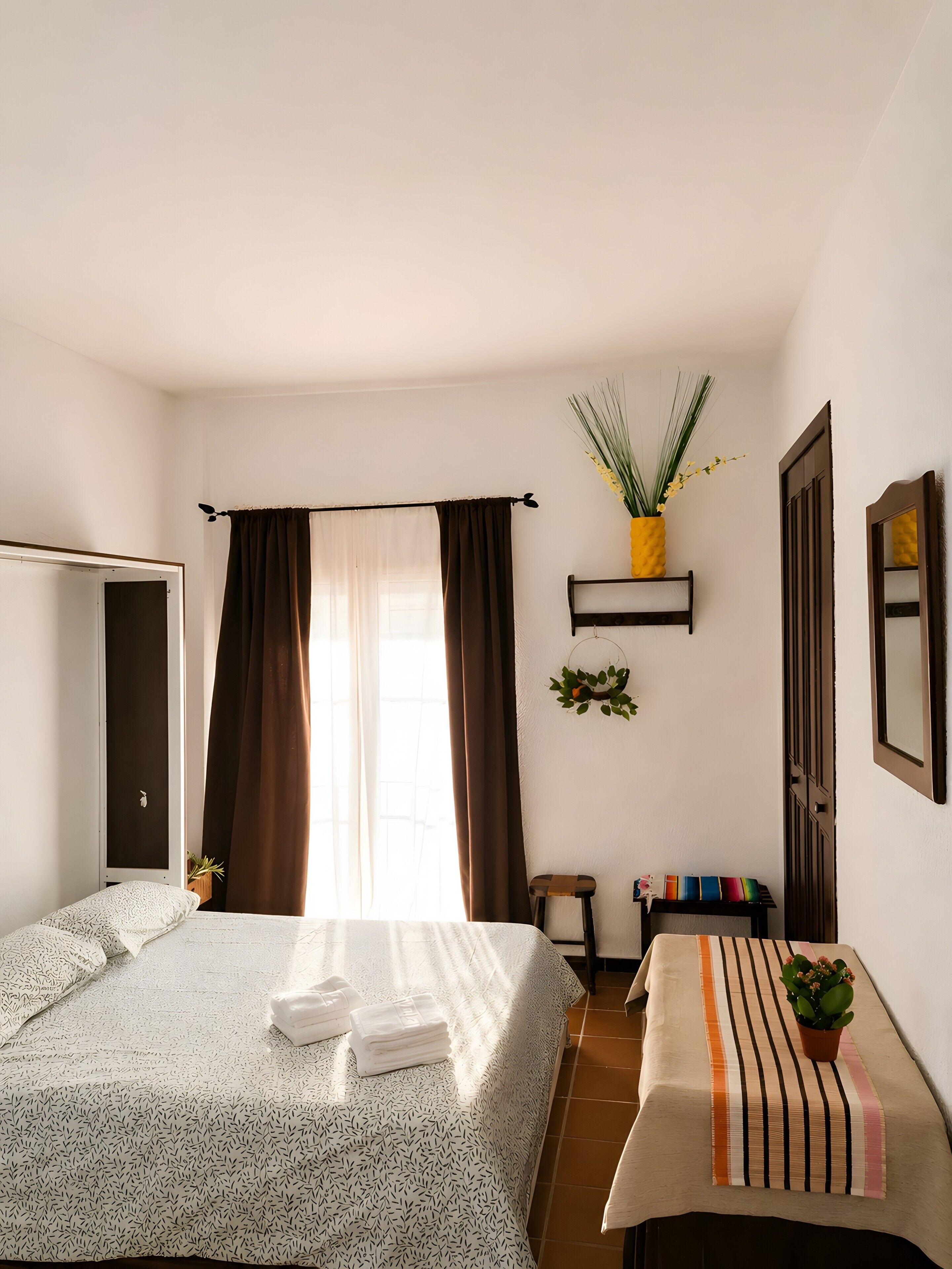 A bedroom with a bed, a bedside table, a chair, a window with curtains, a wall-mounted shelf with a plant, and a door.