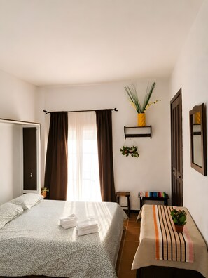 A bedroom with a bed, a bedside table, a chair, a window with curtains, a wall-mounted shelf with a plant, and a door. - Holiday Home 'Acogedor Estudio Junto A La Hermita' with Balcony (El Rocío)