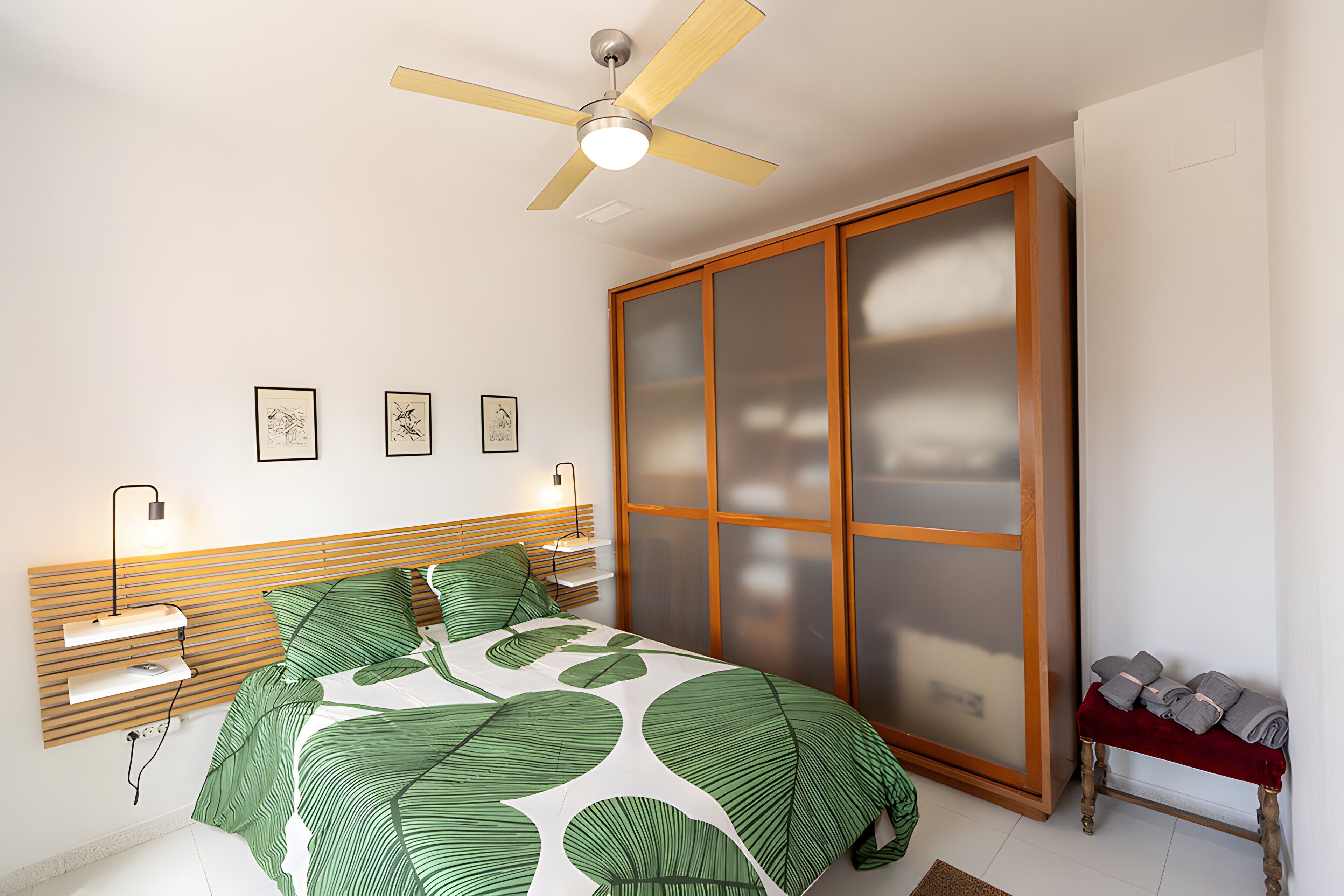 A bedroom with a bed, a ceiling fan, a wardrobe, and wall-mounted lights.