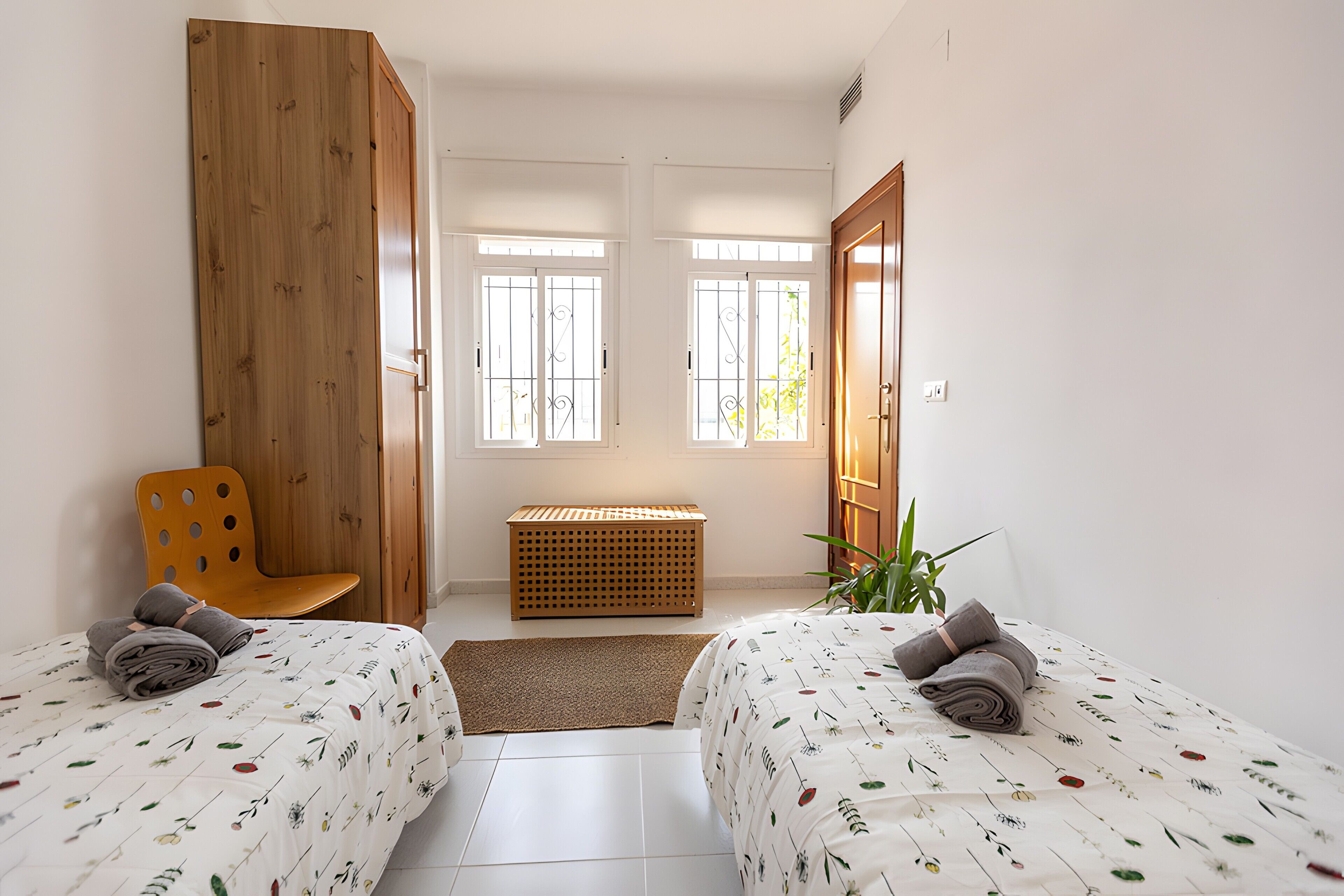 Un dormitorio con dos camas, un armario de madera, un banco, una mesita pequeña y una planta en maceta.