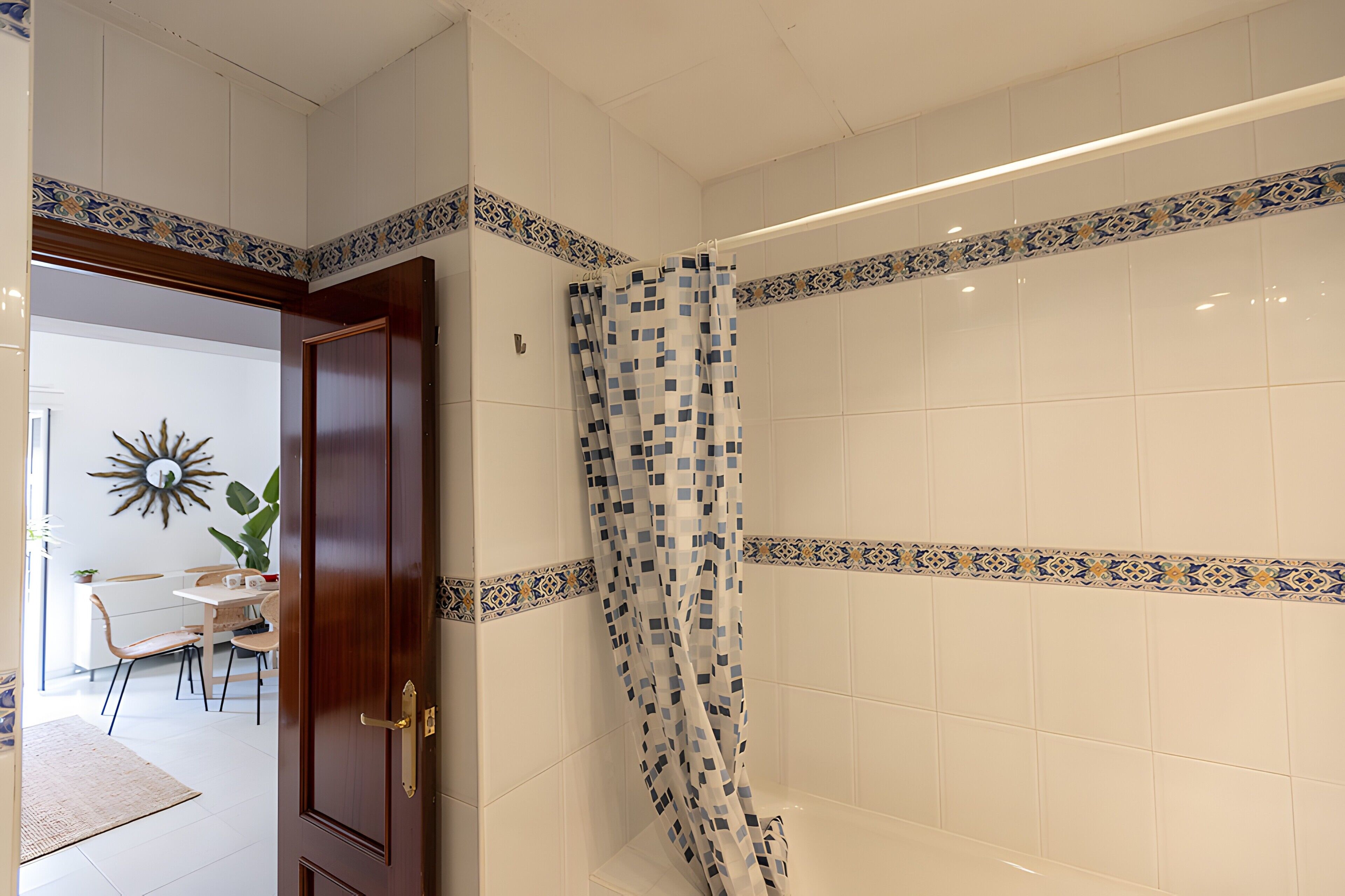 Un baño con azulejos blancos, una cortina de ducha con estampado azul y blanco, y una puerta de madera.