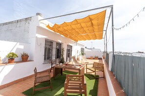 Een terras buiten met een tafel, stoelen, potplanten en een geel zonnescherm.