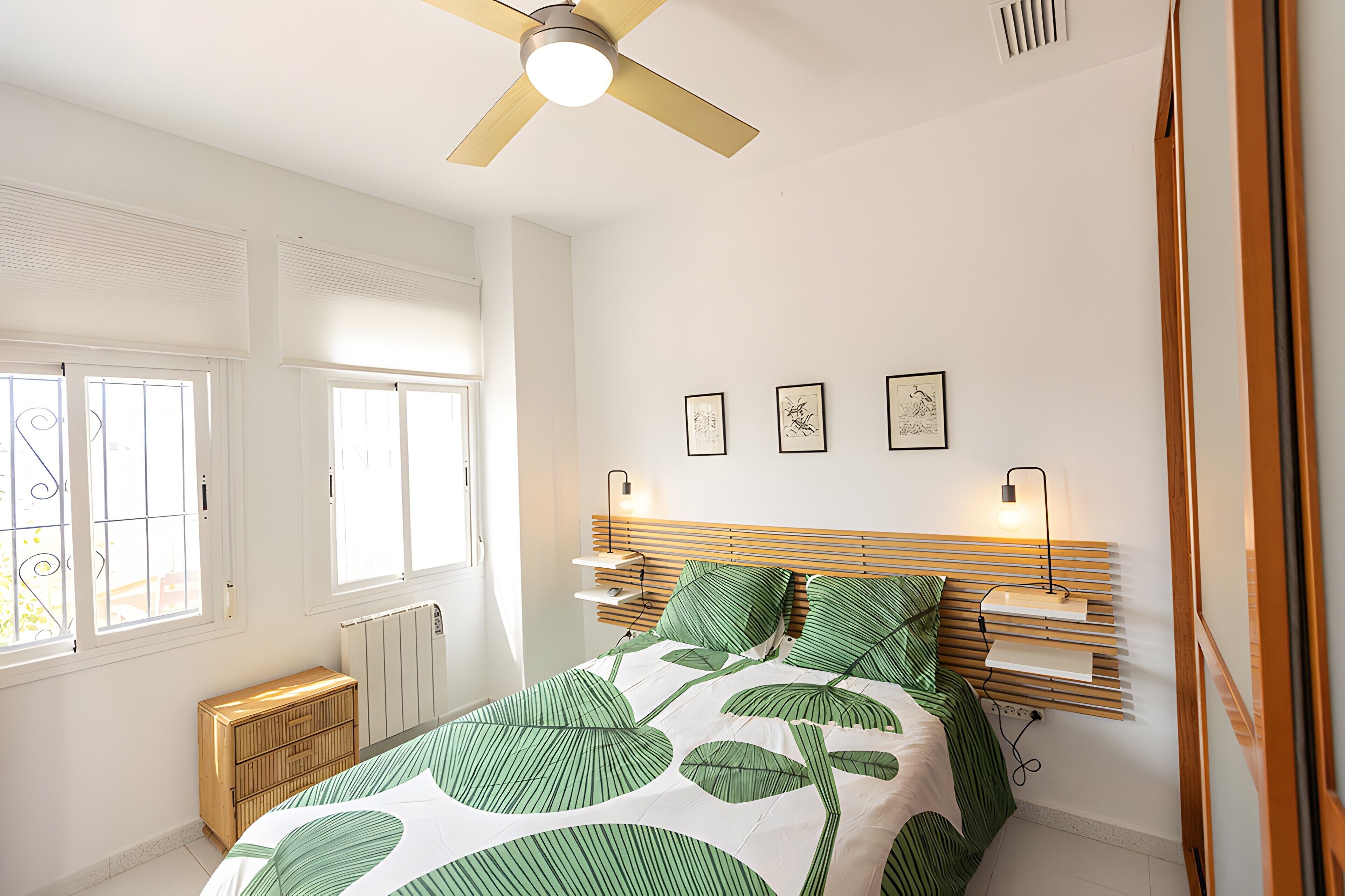 Un dormitorio con una cama, ventilador de techo, radiador, ventana y estantes en la pared.
