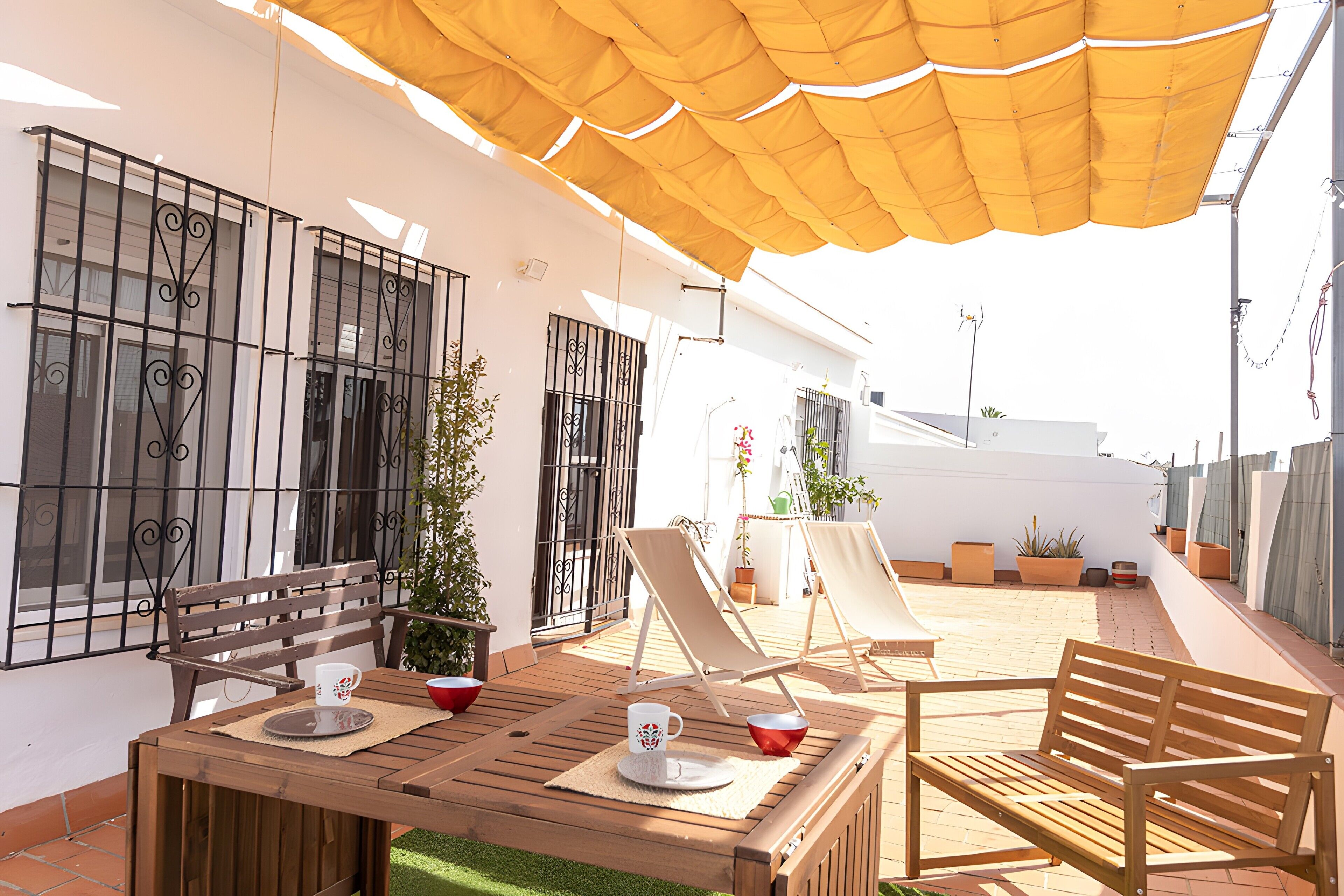 Un patio con una mesa de madera, dos sillas, un banco y una sombrilla amarilla que da sombra.